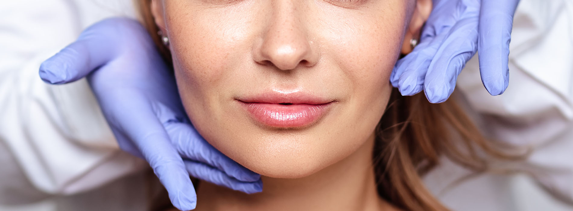 This is a close-up photograph of a person s face with a large magnifying glass over their nose, taken from a perspective that obscures the eyes. The individual appears to be receiving a facial treatment or examination in a professional setting, indicated by the presence of a medical glove and blue surgical drape.