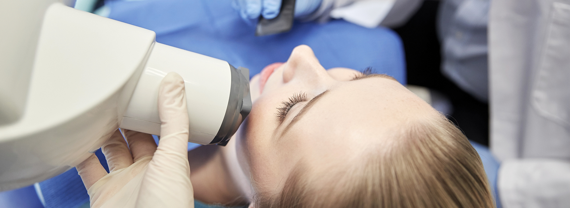 The image shows a person receiving medical attention under a magnifying device, with a healthcare professional closely examining their face using a specialized mirror.