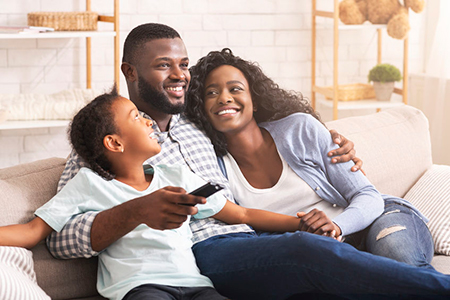 A family of four sitting together on a couch, with a man holding a remote control, smiling at the camera.